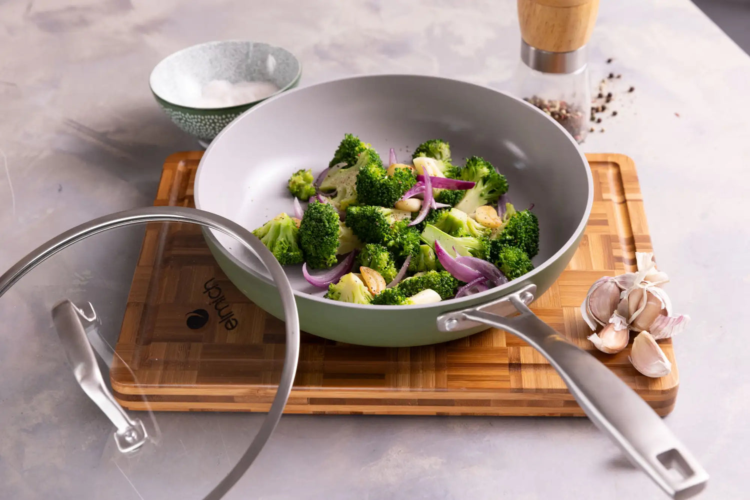 Světle zelená nepřilnavá pánev s nerezovou rukojetí a skleněným víkem, s názvem značky 'anjoy' na spodní straně.